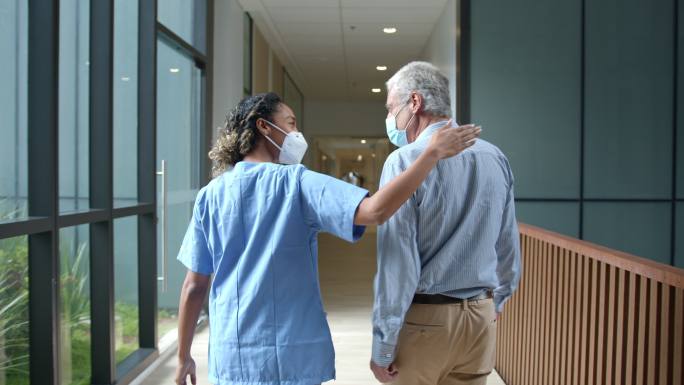 2019冠状病毒疾病大流行期间，一名非裔美国女医生在医院走廊与一名老年患者并肩而行，两人都戴着防护面
