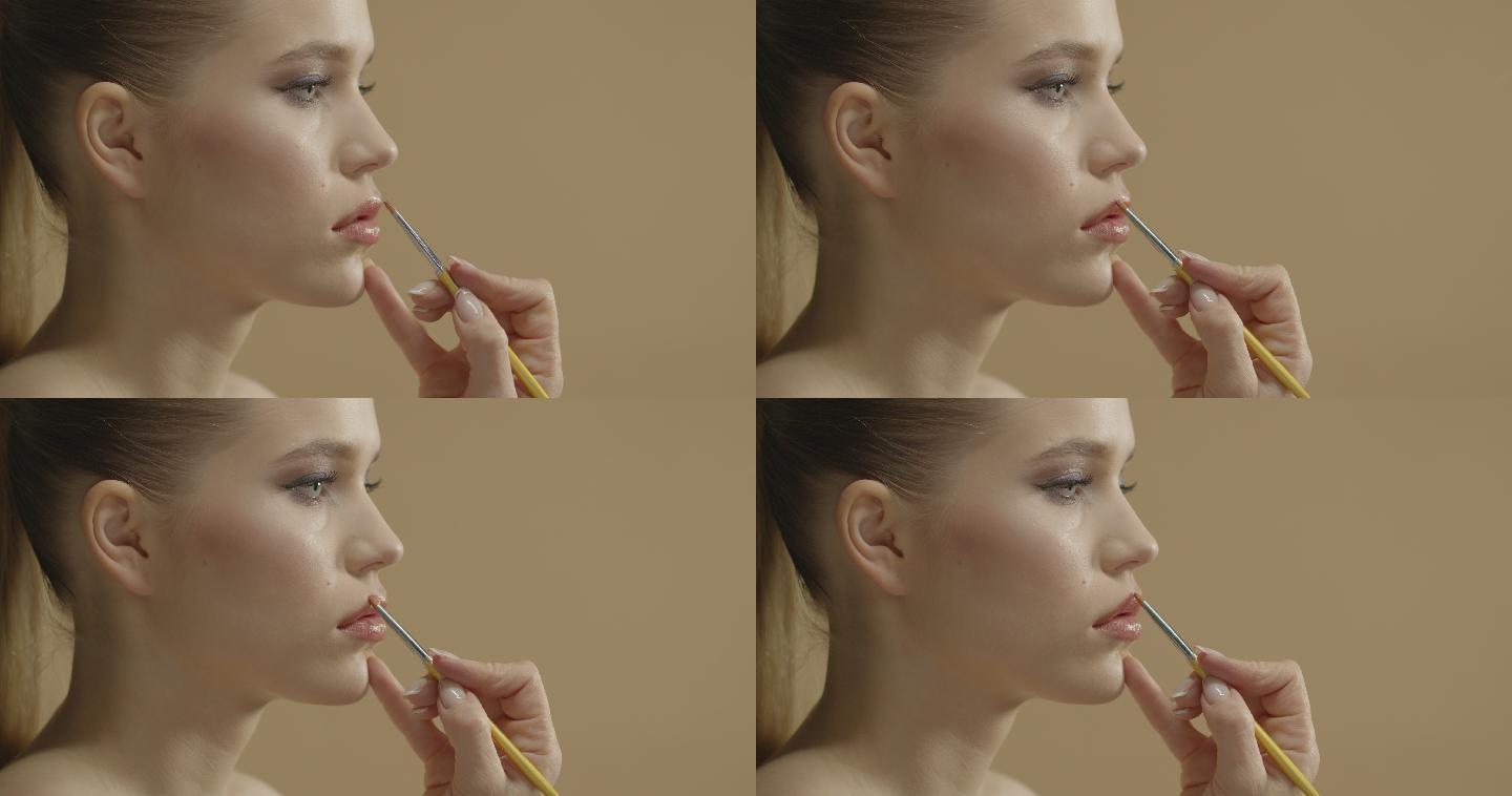 特写镜头中，一位化妆师用眼影刷给一位年轻漂亮的金发女郎化妆，用唇彩涂抹嘴唇。