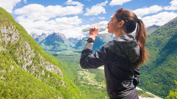 在一个美丽的阳光明媚的日子里，一位女性徒步旅行者一边喝水一边欣赏山景