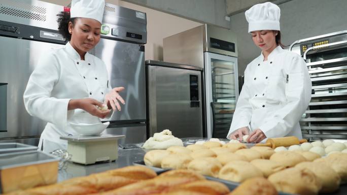 非洲和亚辛年轻的女厨师穿着厨师白，在商业烘焙厨房里一起揉面、称重和准备面包和馒头的面团