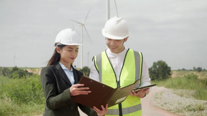 风力发电站的技术工程师。穿着反光夹克和头盔的亚洲男工程师和女商人在风力发电场散步。男子在文件夹中签署