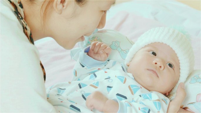 快乐的亚洲母亲在卧室里抱着并亲吻着混血可爱的新生女婴的脸颊