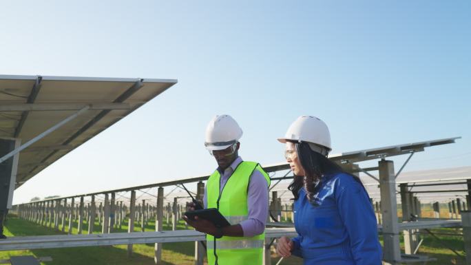 身穿蓝色防护服的亚洲中年女性技术员和穿着反光布的非洲工程师一起工作，使用数字平板电脑检查太阳能发电场