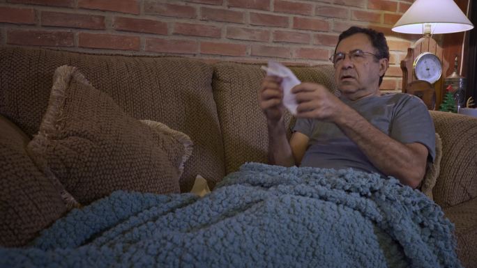 西班牙裔老人躺在沙发上，裹着毯子，可能因新冠肺炎而感到恶心