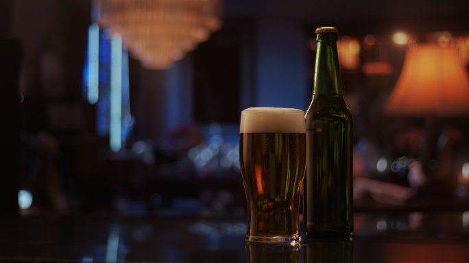 在烛光照亮的黑暗房间里，一杯啤酒和一个玻璃瓶并排放在吧台上