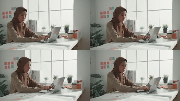 年轻的穆斯林女商人戴着头巾，坐在办公室的办公桌上使用笔记本电脑。