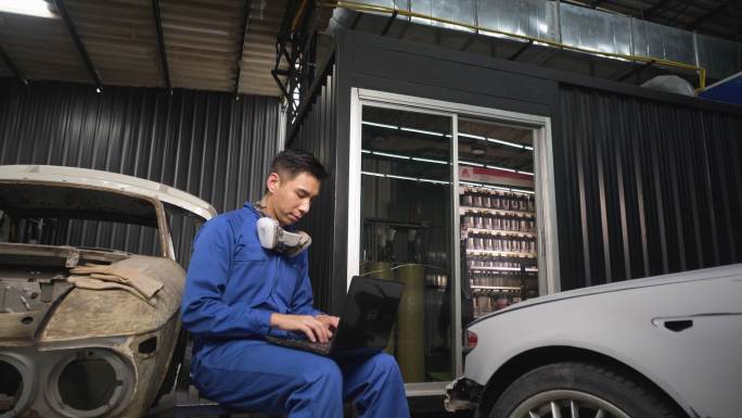 小企业主汽车修理工修理汽车油漆亚洲男性工人在车库工作。使用手机和笔记本电脑进行业务的维修工