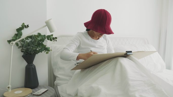 戴着红帽的利基女艺术家正在她的白色卧室里为她的休闲活动进行绘画和素描