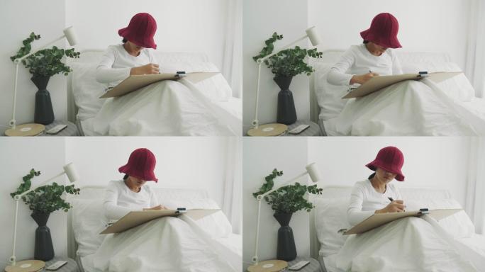戴着红帽的利基女艺术家正在她的白色卧室里为她的休闲活动进行绘画和素描