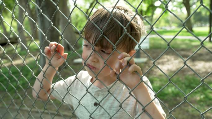 严肃沉思的小男孩透过校园的铁丝网围栏看