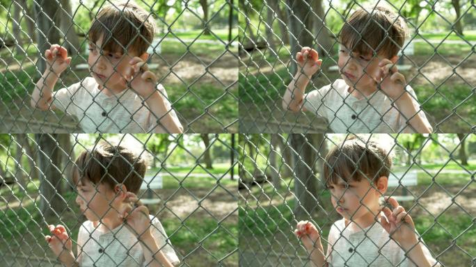 严肃沉思的小男孩透过校园的铁丝网围栏看