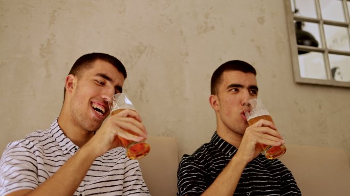 同卵双胞胎兄弟坐着，微笑着，喝啤酒，使用智能手机