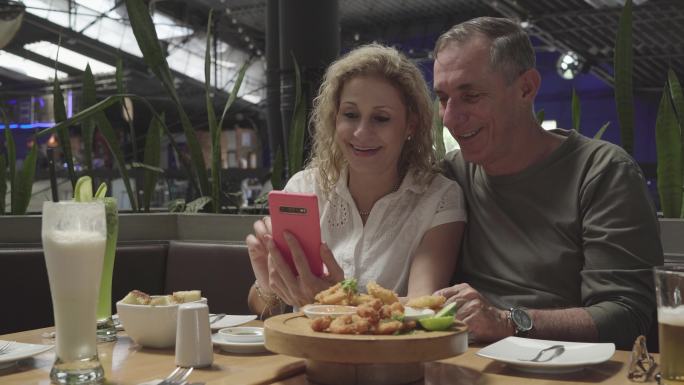 一对年纪较大的拉丁夫妇在一家餐厅里享受着他们的情侣时光，看着手机上拍摄的照片