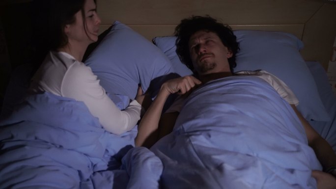 一对异性伴侣躺在床上，男人睡觉时张嘴打鼾，而一个疲惫的女人则头枕枕头坐在床上打鼾。