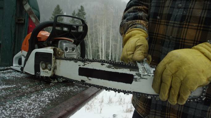 一位30多岁留着胡子的白人男子在森林里的卡车尾门上磨着电锯，这是一个大雪纷飞的冬日，在山里