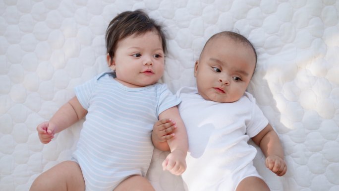 头顶上拍摄的是两个穿着白色婴儿服的快乐无辜的黑白新生儿躺在柔软的床上，双臂交叉在一起玩耍