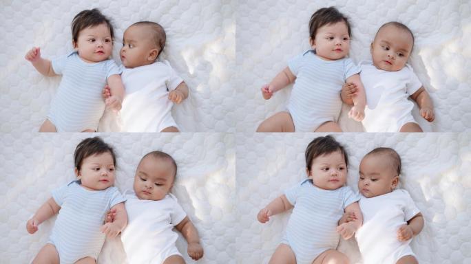头顶上拍摄的是两个穿着白色婴儿服的快乐无辜的黑白新生儿躺在柔软的床上，双臂交叉在一起玩耍