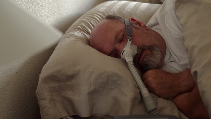患有睡眠呼吸暂停症的中年男子躺在床上睡觉，他戴着CPAP（持续气道正压通气）机器帮助自己入睡