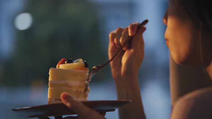 日落时分，中国亚裔美女在家吃蛋糕，透过窗帘远眺