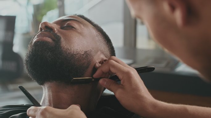 一个英俊的年轻人坐在理发店里，被理发师剃胡子的4k视频片段
