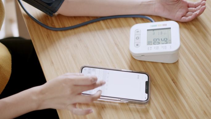 POV：亚洲女性在血压监护仪上自我测量血压和心率，在家中使用智能手机记录数据