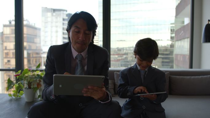 父亲和儿子都穿着工作服坐在沙发上，各自在平板电脑上工作