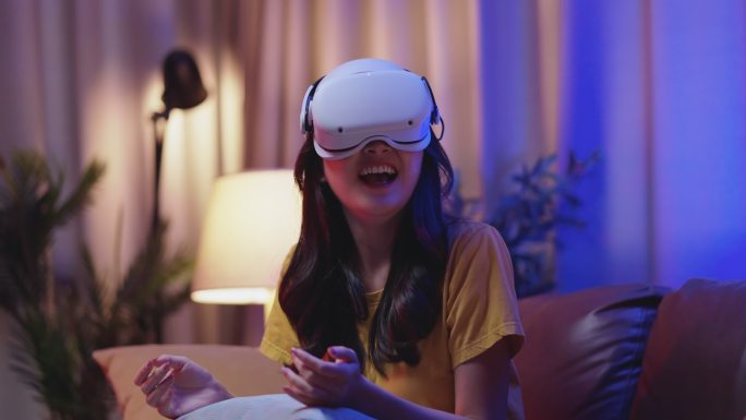 年轻迷人的休闲亚裔女性在家里的客厅里，通过VR眼镜、耳机、护目镜，在沙发上观看虚拟现实中的表演生活流