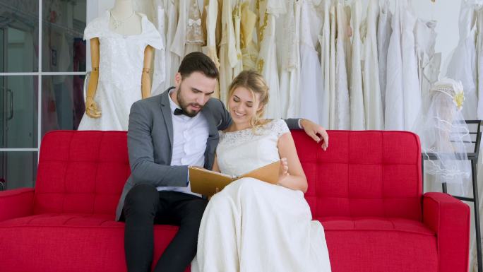 一对年轻的休闲情侣坐在婚纱店的沙发上看书，挑选最佳婚纱。