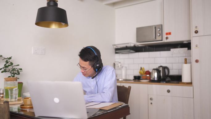 亚裔中国中年男子在用餐区使用笔记本电脑和耳机微笑着参加在线会议