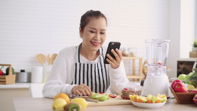 亚洲资深女性一边吃水果一边打电话，准备自制不同口味的水果冰沙和酸奶，感觉很幸福。健康天然益生菌口味饮