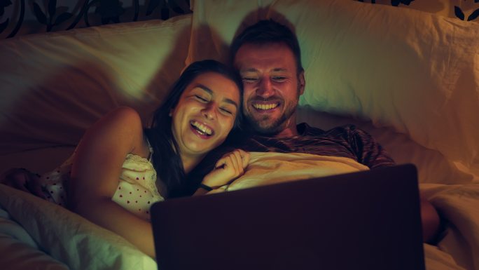 这对年轻夫妇在床上看电影。使用笔记本电脑，享受乐趣