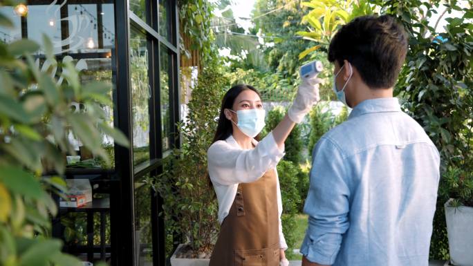亚洲女性咖啡馆员工在服用体温后，亚洲男性在前往餐厅或咖啡馆前戴上口罩，以适应新的正常生活方式冠状病毒