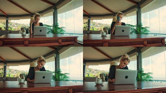 在全景窗旁工作的妇女。使用笔记本电脑和咖啡欣赏史诗般的海景