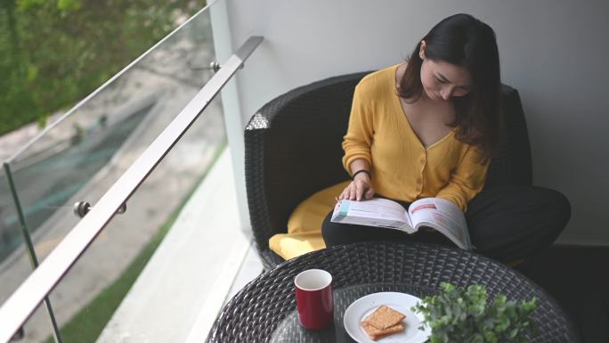 一位亚裔华裔美女中午在阳台上看书，放松身心，享受着用咖啡和饼干作为零食的数字排毒时光
