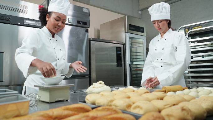 在商业面包房厨房，身着厨师白的非洲和亚细亚年轻女厨师一起帮助揉捏、称量和准备面团，制作面包和馒头