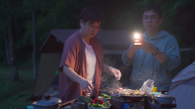 一名亚裔中国女子晚上在野营时准备烤肉，而她的丈夫则用智能手机点亮了现场