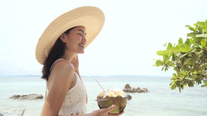 美丽的亚洲年轻女子在海滩上喝着椰子水。