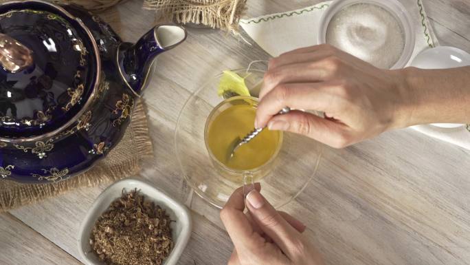 一张白色乡村桌子上，一位女士双手向热茶甘菊输液杯中倒入和搅拌一茶匙糖的高角度特写照片