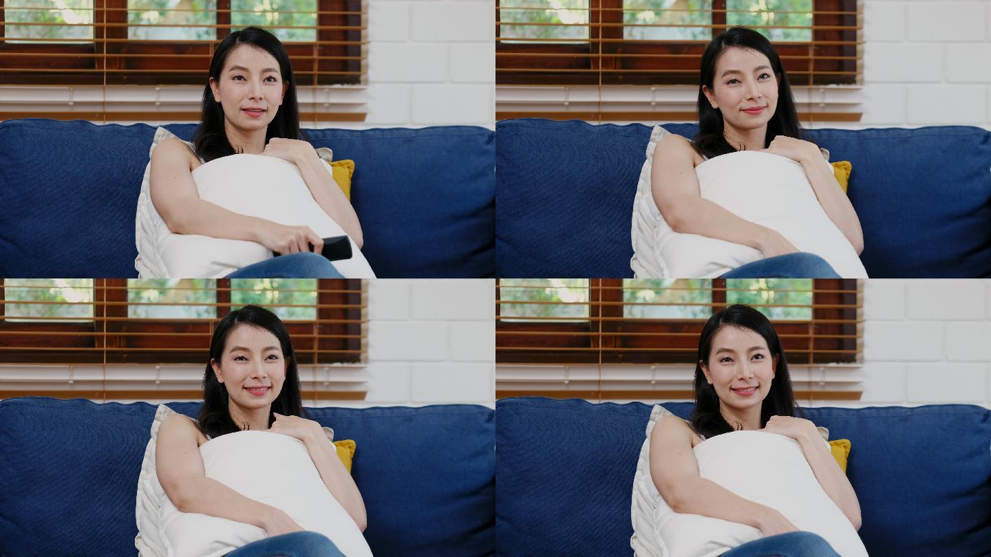 年轻的亚洲女性一边看电视一边拿着遥控器坐在家客厅背景的沙发上，人们享受着生活方式