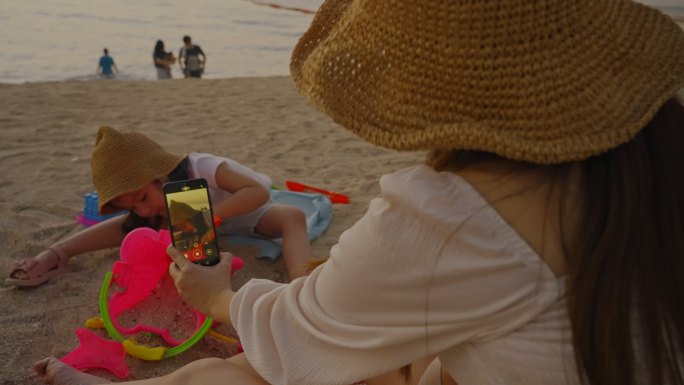 母亲用智能手机拍照，孩子在沙滩上玩沙堡