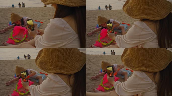 母亲用智能手机拍照，孩子在沙滩上玩沙堡