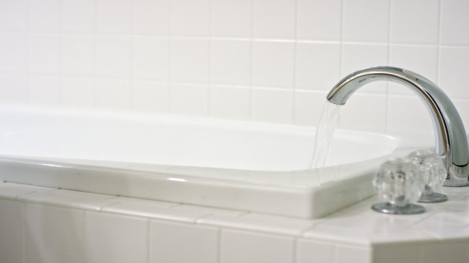 一位白人妇女走到一个铺着白色瓷砖的浴缸前，打开水龙头洗澡