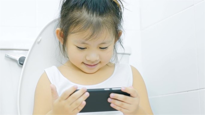 亚洲小女孩看着智能手机，坐在浴室的马桶上