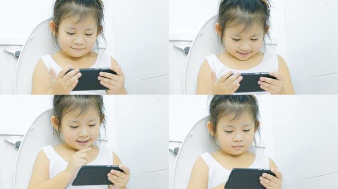亚洲小女孩看着智能手机，坐在浴室的马桶上
