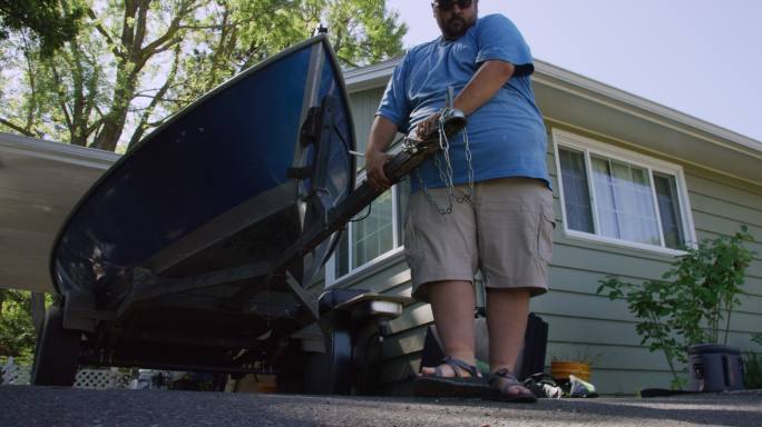 一名40多岁的西班牙裔男子将拖车上的一艘小船挂在汽车的挂接装置上，以便在阳光明媚的日子将其拖到屋外