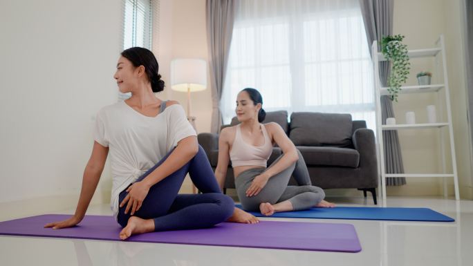 两位苗条的亚洲女性正在做半鱼王瑜伽姿势，右膝弯曲，右脚缩进左臀，右臂在右耳旁笔直向上，然后将躯干向左