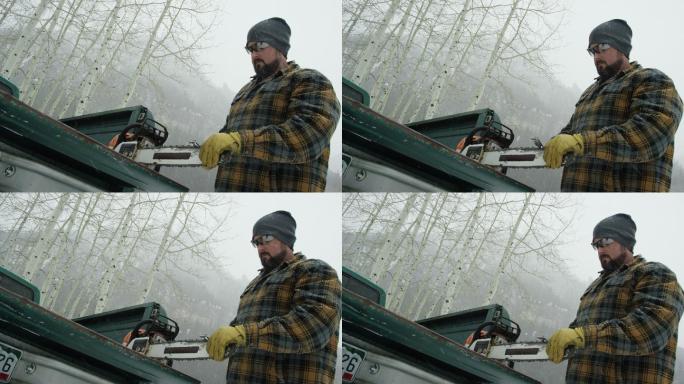 一位30多岁留着胡子的白人男子在森林里的卡车尾门上磨着电锯，这是一个大雪纷飞的冬日，在山里（中景）
