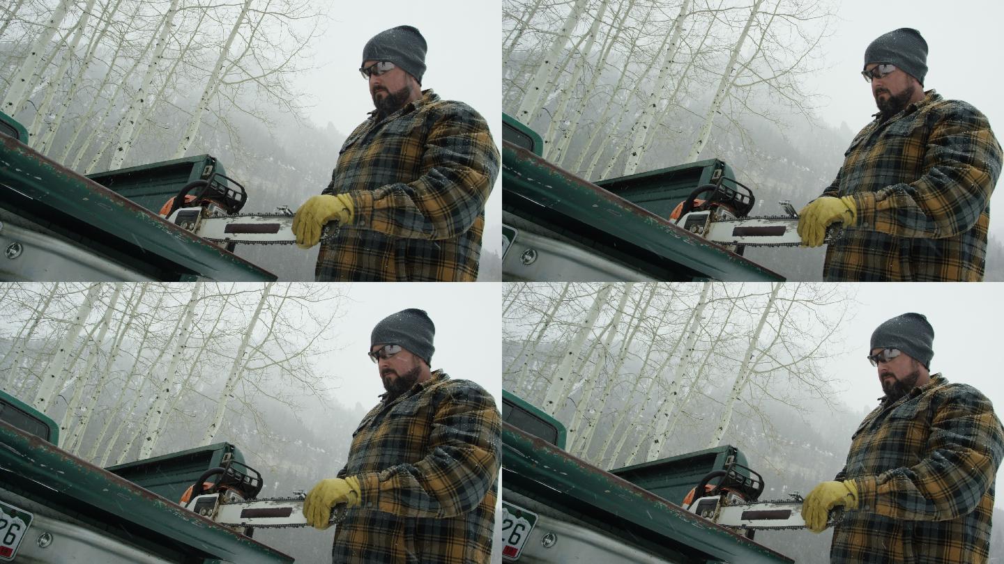 一位30多岁留着胡子的白人男子在森林里的卡车尾门上磨着电锯，这是一个大雪纷飞的冬日，在山里（中景）
