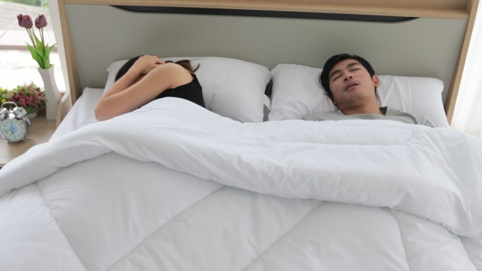 亚洲年轻的妻子睡不着，丈夫在她身边打鼾，女人用枕头捂住耳朵。