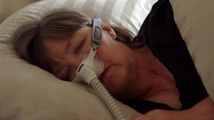患有睡眠呼吸暂停症的中年女性在床上睡觉时，戴着CPAP（持续气道正压通气）机器帮助睡眠